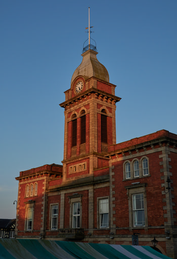 The image is an architecture photograph taken in the morning, showing the Chesterfield Market Hall in Chesterfield. The Market Hall is captured in early summer, evident from the clear blue sky and soft morning sunlight illuminating the red brickwork and stone detailing of the historic building. The prominent clock tower, topped with a weather vane and flagpole, stands out as the main subject, and is a well-known landmark within the town. Large sash windows and classical design elements around the Market Hall emphasize its architectural significance in the heart of Chesterfield. The photograph demonstrates the building’s distinctive features, with gentle sunlight creating warm highlights typical of a summer morning.